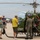 A group of people gather around a vintage U.S. Army helicopter at what appears to be an outdoor airshow or aviation exhibition. The helicopter is olive green with 'ARMY' lettering and a U.S. military star insignia, with its engine compartment exposed for viewing. A person in a flight suit appears to be explaining or demonstrating the aircraft to interested spectators.