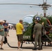 A group of people gather around a vintage U.S. Army helicopter at what appears to be an outdoor airshow or aviation exhibition. The helicopter is olive green with 'ARMY' lettering and a U.S. military star insignia, with its engine compartment exposed for viewing. A person in a flight suit appears to be explaining or demonstrating the aircraft to interested spectators.