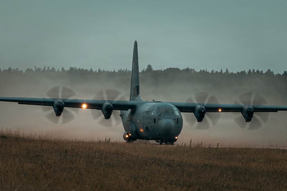 A military C-130 Hercules turboprop transport aircraft is taxiing or landing on a dirt/grass airstrip under overcast, low-light conditions. The four propellers are spinning at high speed, creating visible dust clouds and prop wash behind the aircraft. The text 'AIR FORCE' is partially visible on the fuselage, indicating a US Air Force aircraft.