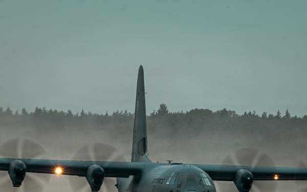 A military C-130 Hercules turboprop transport aircraft is taxiing or landing on a dirt/grass airstrip under overcast, low-light conditions. The four propellers are spinning at high speed, creating visible dust clouds and prop wash behind the aircraft. The text 'AIR FORCE' is partially visible on the fuselage, indicating a US Air Force aircraft.