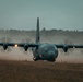 A military C-130 Hercules turboprop transport aircraft is taxiing or landing on a dirt/grass airstrip under overcast, low-light conditions. The four propellers are spinning at high speed, creating visible dust clouds and prop wash behind the aircraft. The text 'AIR FORCE' is partially visible on the fuselage, indicating a US Air Force aircraft.