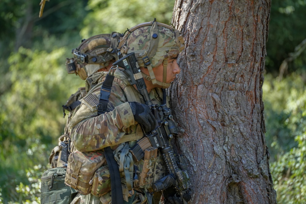 Two heavily equipped soldiers in multicam/OCP camouflage uniforms are taking cover behind a large tree trunk in a wooded environment. The foreground soldier is peering around the tree while holding an assault rifle, suggesting a tactical movement or reconnaissance exercise. Both soldiers are wearing full combat gear including helmets, plate carriers, and tactical accessories.