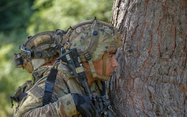 Two heavily equipped soldiers in multicam/OCP camouflage uniforms are taking cover behind a large tree trunk in a wooded environment. The foreground soldier is peering around the tree while holding an assault rifle, suggesting a tactical movement or reconnaissance exercise. Both soldiers are wearing full combat gear including helmets, plate carriers, and tactical accessories.