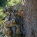 Two heavily equipped soldiers in multicam/OCP camouflage uniforms are taking cover behind a large tree trunk in a wooded environment. The foreground soldier is peering around the tree while holding an assault rifle, suggesting a tactical movement or reconnaissance exercise. Both soldiers are wearing full combat gear including helmets, plate carriers, and tactical accessories.