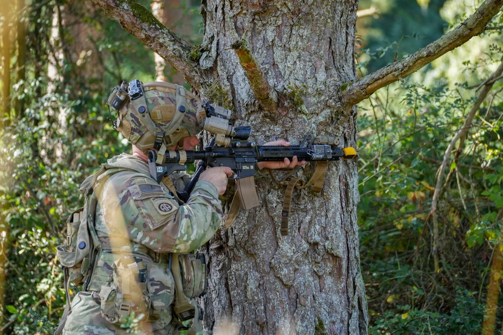 A U.S. Army soldier in full combat gear is taking a firing position behind a large tree in a forested environment. The soldier is aiming a rifle equipped with optics and accessories, using the tree as cover. Visible unit patches and equipment suggest this is a training exercise rather than active combat.