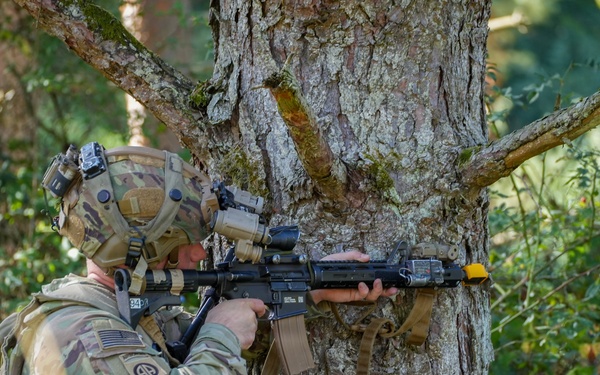 A U.S. Army soldier in full combat gear is taking a firing position behind a large tree in a forested environment. The soldier is aiming a rifle equipped with optics and accessories, using the tree as cover. Visible unit patches and equipment suggest this is a training exercise rather than active combat.