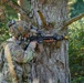 A U.S. Army soldier in full combat gear is taking a firing position behind a large tree in a forested environment. The soldier is aiming a rifle equipped with optics and accessories, using the tree as cover. Visible unit patches and equipment suggest this is a training exercise rather than active combat.