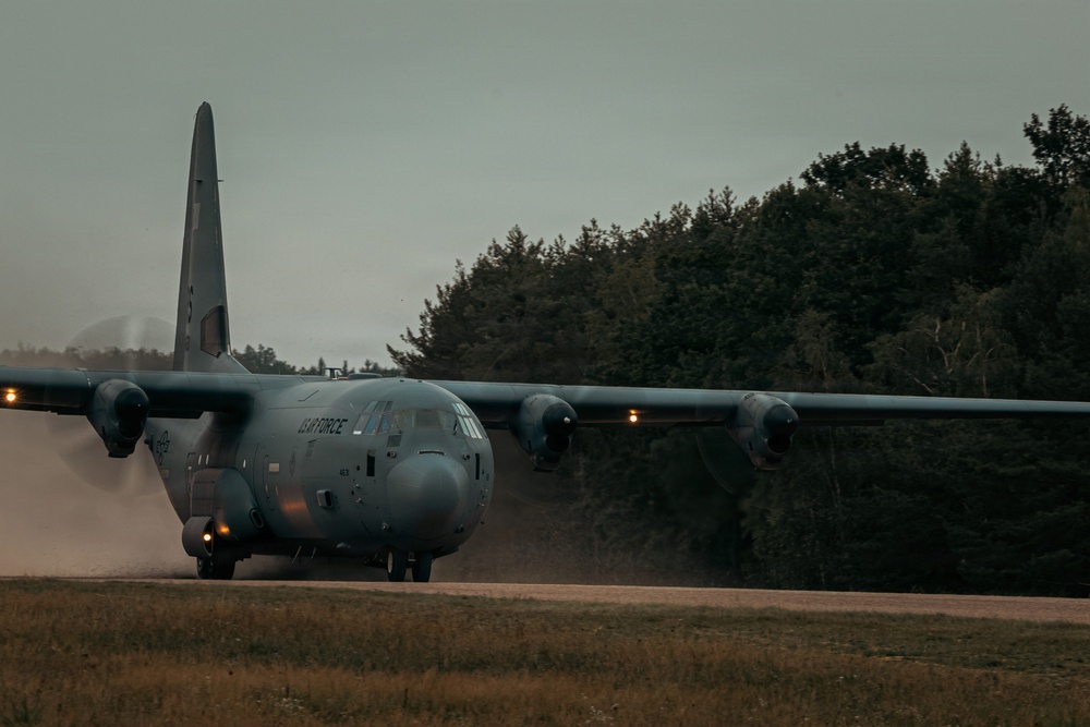 A U.S. Air Force C-130 Hercules military transport aircraft is operating on what appears to be an unpaved or semi-prepared airstrip, kicking up dust and debris from its turboprop engines. The aircraft is marked with 'US AIR FORCE' on the fuselage and is photographed in low-light, overcast conditions. A dense treeline forms the background, suggesting a remote or austere landing zone environment.