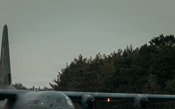 A U.S. Air Force C-130 Hercules military transport aircraft is operating on what appears to be an unpaved or semi-prepared airstrip, kicking up dust and debris from its turboprop engines. The aircraft is marked with 'US AIR FORCE' on the fuselage and is photographed in low-light, overcast conditions. A dense treeline forms the background, suggesting a remote or austere landing zone environment.