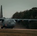 A U.S. Air Force C-130 Hercules military transport aircraft is operating on what appears to be an unpaved or semi-prepared airstrip, kicking up dust and debris from its turboprop engines. The aircraft is marked with 'US AIR FORCE' on the fuselage and is photographed in low-light, overcast conditions. A dense treeline forms the background, suggesting a remote or austere landing zone environment.