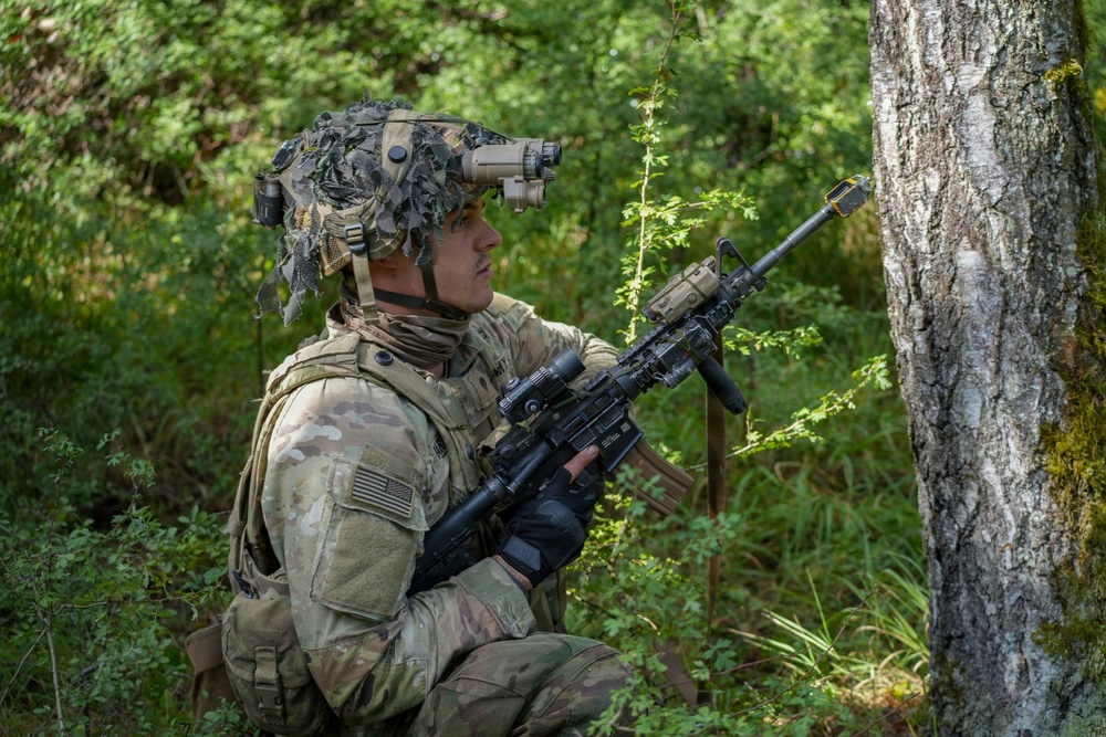 A U.S. Army soldier in full combat gear is positioned behind a tree in a forested environment, appearing to conduct a reconnaissance or patrol exercise. The soldier is equipped with multicam camouflage, a helmet-mounted night vision device, and an M4-style carbine with accessories. The setting suggests a training exercise or field operation in a temperate woodland environment.