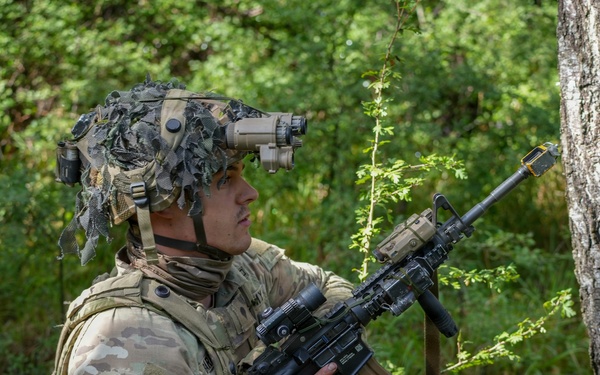 A U.S. Army soldier in full combat gear is positioned behind a tree in a forested environment, appearing to conduct a reconnaissance or patrol exercise. The soldier is equipped with multicam camouflage, a helmet-mounted night vision device, and an M4-style carbine with accessories. The setting suggests a training exercise or field operation in a temperate woodland environment.