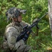 A U.S. Army soldier in full combat gear is positioned behind a tree in a forested environment, appearing to conduct a reconnaissance or patrol exercise. The soldier is equipped with multicam camouflage, a helmet-mounted night vision device, and an M4-style carbine with accessories. The setting suggests a training exercise or field operation in a temperate woodland environment.