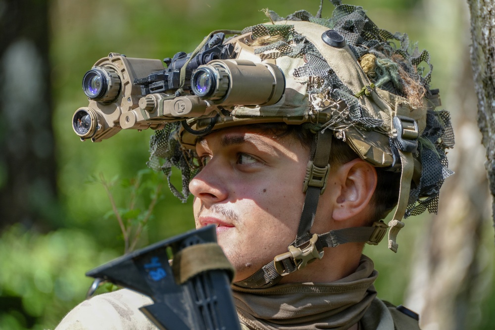 A young male soldier is photographed in a forested outdoor environment wearing a camouflaged tactical helmet equipped with a quad-tube night vision goggle (NVG) system. The soldier appears alert and is looking upward or to the side, suggesting situational awareness during a field exercise or operation. The image is a close-up portrait-style shot with high detail on the equipment and facial features.