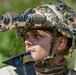 A young male soldier is photographed in a forested outdoor environment wearing a camouflaged tactical helmet equipped with a quad-tube night vision goggle (NVG) system. The soldier appears alert and is looking upward or to the side, suggesting situational awareness during a field exercise or operation. The image is a close-up portrait-style shot with high detail on the equipment and facial features.