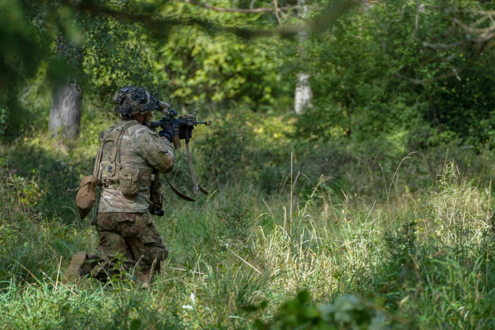 A soldier in full combat gear is kneeling in a wooded, overgrown area while aiming a rifle forward. The soldier is wearing camouflage uniform, tactical vest, helmet with attachments, and carrying a backpack. The setting appears to be a training exercise or patrol in a dense green forest environment.