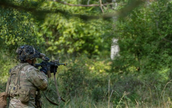 A soldier in full combat gear is kneeling in a wooded, overgrown area while aiming a rifle forward. The soldier is wearing camouflage uniform, tactical vest, helmet with attachments, and carrying a backpack. The setting appears to be a training exercise or patrol in a dense green forest environment.