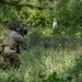 A soldier in full combat gear is kneeling in a wooded, overgrown area while aiming a rifle forward. The soldier is wearing camouflage uniform, tactical vest, helmet with attachments, and carrying a backpack. The setting appears to be a training exercise or patrol in a dense green forest environment.