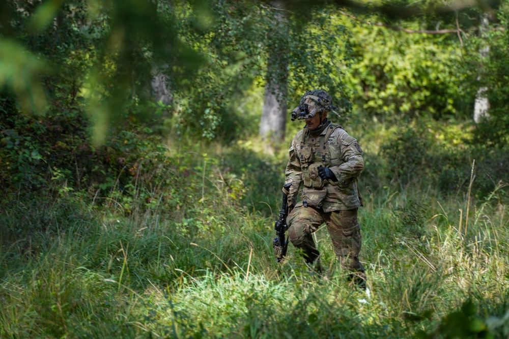 A fully equipped soldier in multicam/OCP camouflage uniform is moving through a dense woodland or forest clearing with tall grass and green vegetation. The soldier is carrying a weapon and wearing full combat gear including a helmet with a mounted device, body armor, and tactical accessories. The setting appears to be a field training exercise or operational patrol in a temperate forested environment.