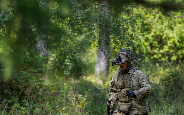 A fully equipped soldier in multicam/OCP camouflage uniform is moving through a dense woodland or forest clearing with tall grass and green vegetation. The soldier is carrying a weapon and wearing full combat gear including a helmet with a mounted device, body armor, and tactical accessories. The setting appears to be a field training exercise or operational patrol in a temperate forested environment.