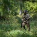 A fully equipped soldier in multicam/OCP camouflage uniform is moving through a dense woodland or forest clearing with tall grass and green vegetation. The soldier is carrying a weapon and wearing full combat gear including a helmet with a mounted device, body armor, and tactical accessories. The setting appears to be a field training exercise or operational patrol in a temperate forested environment.