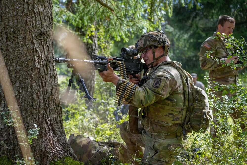 A U.S. Army soldier in Operational Camouflage Pattern (OCP) uniform is firing a belt-fed machine gun, likely an M249 SAW or M240, from a kneeling position near a tree in a wooded area. Visible muzzle smoke and a linked ammunition belt indicate active firing. A second soldier in the background appears to be observing or providing support, suggesting a training exercise or field operation.