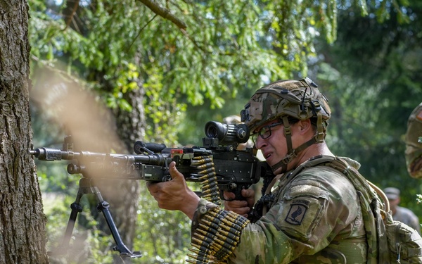 A U.S. Army soldier in Operational Camouflage Pattern (OCP) uniform is firing a belt-fed machine gun, likely an M249 SAW or M240, from a kneeling position near a tree in a wooded area. Visible muzzle smoke and a linked ammunition belt indicate active firing. A second soldier in the background appears to be observing or providing support, suggesting a training exercise or field operation.