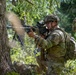 A U.S. Army soldier in Operational Camouflage Pattern (OCP) uniform is firing a belt-fed machine gun, likely an M249 SAW or M240, from a kneeling position near a tree in a wooded area. Visible muzzle smoke and a linked ammunition belt indicate active firing. A second soldier in the background appears to be observing or providing support, suggesting a training exercise or field operation.