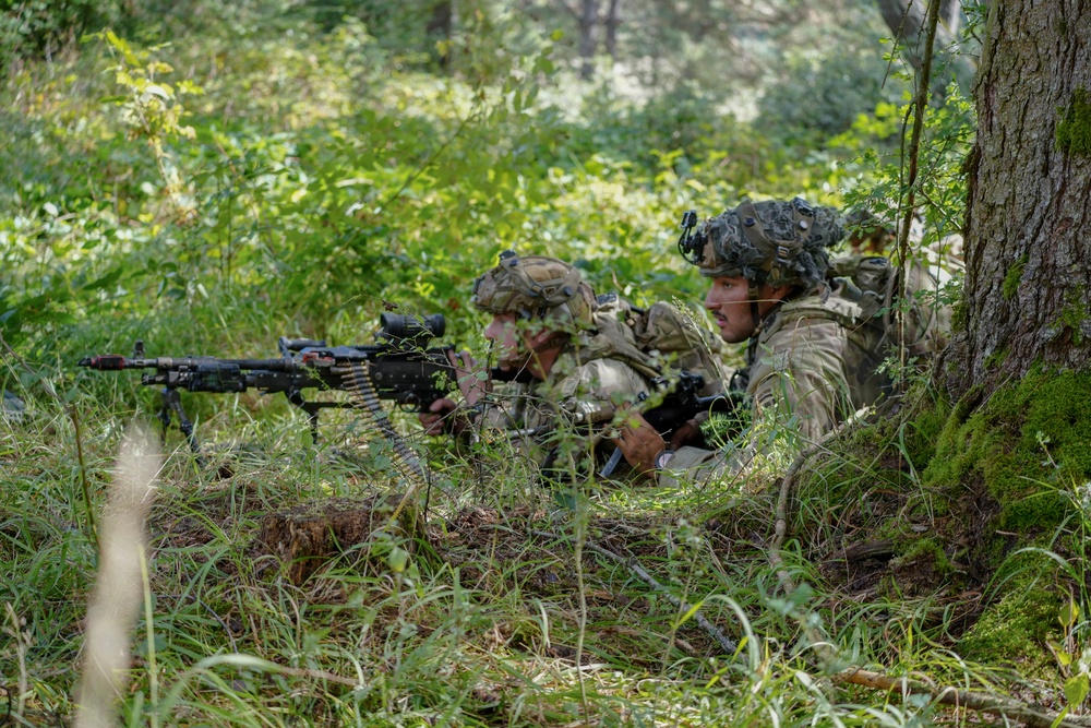 Two soldiers in full combat gear are positioned prone at the base of a large tree in a forested environment, taking up a defensive or observation position. The soldier on the left is manning what appears to be a belt-fed machine gun aimed forward, while the soldier on the right observes the surroundings. The setting and equipment suggest a military training exercise or field operation.