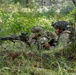 Two soldiers in full combat gear are positioned prone at the base of a large tree in a forested environment, taking up a defensive or observation position. The soldier on the left is manning what appears to be a belt-fed machine gun aimed forward, while the soldier on the right observes the surroundings. The setting and equipment suggest a military training exercise or field operation.