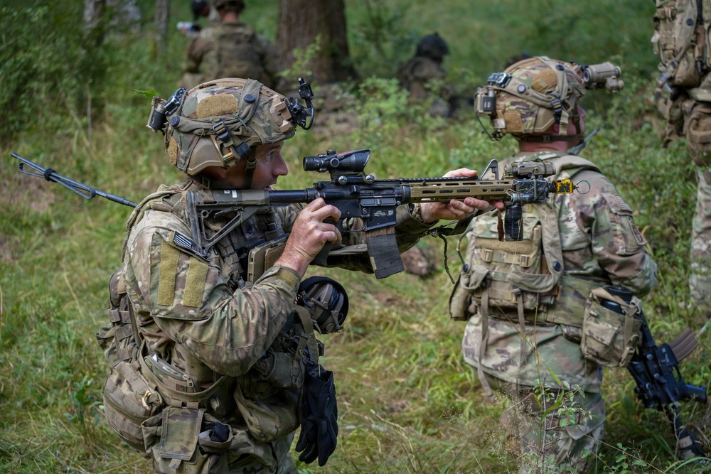 The image shows multiple soldiers in full combat gear conducting what appears to be a military training exercise or patrol in a wooded, vegetated area. The foreground soldier is in a kneeling position aiming an assault rifle equipped with an optic scope, while a second soldier stands nearby. Additional personnel are visible in the background among the trees.