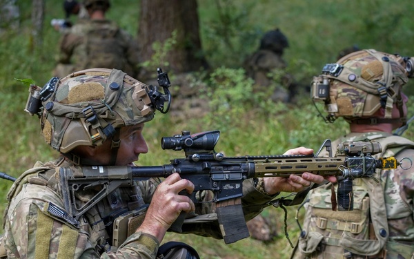 The image shows multiple soldiers in full combat gear conducting what appears to be a military training exercise or patrol in a wooded, vegetated area. The foreground soldier is in a kneeling position aiming an assault rifle equipped with an optic scope, while a second soldier stands nearby. Additional personnel are visible in the background among the trees.