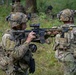 The image shows multiple soldiers in full combat gear conducting what appears to be a military training exercise or patrol in a wooded, vegetated area. The foreground soldier is in a kneeling position aiming an assault rifle equipped with an optic scope, while a second soldier stands nearby. Additional personnel are visible in the background among the trees.