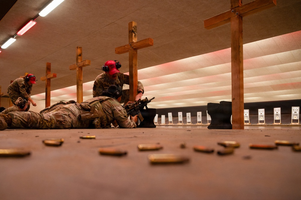 An indoor shooting range training session showing military or law enforcement personnel in camouflage uniforms practicing prone shooting positions. Instructors wearing red caps and hearing protection are supervising a shooter lying on the floor with a rifle. Numerous spent brass casings are visible on the floor in the foreground.