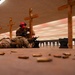 An indoor shooting range training session showing military or law enforcement personnel in camouflage uniforms practicing prone shooting positions. Instructors wearing red caps and hearing protection are supervising a shooter lying on the floor with a rifle. Numerous spent brass casings are visible on the floor in the foreground.