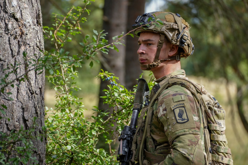 A U.S. Army soldier in full combat gear stands alert in a wooded environment, partially concealed behind a tree and vegetation. The soldier wears MultiCam (OCP) camouflage and displays a 173rd Airborne Brigade Combat Team patch with an Airborne tab on the left shoulder. The image appears to be taken during a field training exercise or operational deployment in a European forest setting.