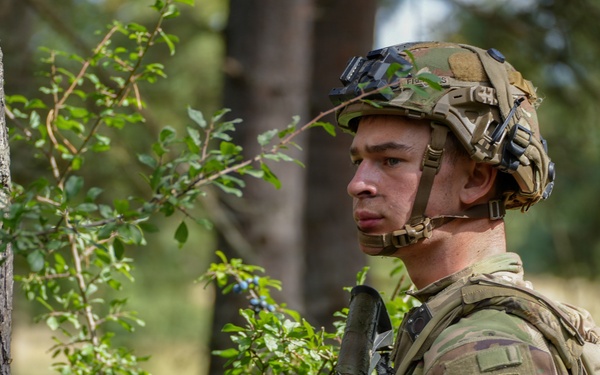 A U.S. Army soldier in full combat gear stands alert in a wooded environment, partially concealed behind a tree and vegetation. The soldier wears MultiCam (OCP) camouflage and displays a 173rd Airborne Brigade Combat Team patch with an Airborne tab on the left shoulder. The image appears to be taken during a field training exercise or operational deployment in a European forest setting.