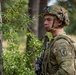 A U.S. Army soldier in full combat gear stands alert in a wooded environment, partially concealed behind a tree and vegetation. The soldier wears MultiCam (OCP) camouflage and displays a 173rd Airborne Brigade Combat Team patch with an Airborne tab on the left shoulder. The image appears to be taken during a field training exercise or operational deployment in a European forest setting.