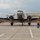 A vintage twin-engine propeller aircraft, consistent with a Douglas DC-3 or similar era transport plane, is taxiing on a concrete airport ramp with both engines running. A ground crew member in a high-visibility orange safety vest is using marshalling wands to guide the aircraft. The setting appears to be an airshow or aviation event, evidenced by crowd barriers and spectators visible in the background.