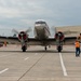 A vintage twin-engine propeller aircraft, consistent with a Douglas DC-3 or similar era transport plane, is taxiing on a concrete airport ramp with both engines running. A ground crew member in a high-visibility orange safety vest is using marshalling wands to guide the aircraft. The setting appears to be an airshow or aviation event, evidenced by crowd barriers and spectators visible in the background.