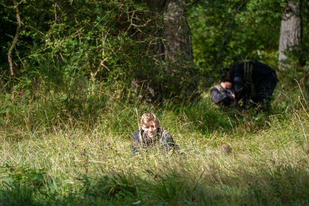 The image shows two individuals in a woodland or forest clearing with tall grass and dense green vegetation. One person, appearing to be a young blonde individual, is lying or crawling low in the grass in the foreground and appears to be smiling. A second person dressed in dark clothing is crouched or bent over in the background near the tree line.
