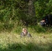 The image shows two individuals in a woodland or forest clearing with tall grass and dense green vegetation. One person, appearing to be a young blonde individual, is lying or crawling low in the grass in the foreground and appears to be smiling. A second person dressed in dark clothing is crouched or bent over in the background near the tree line.
