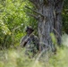 A soldier in full camouflage uniform and tactical helmet is positioned behind a large tree trunk in a dense woodland environment. The soldier is holding a rifle and appears to be in a tactical observation or patrol posture. The image captures a military field exercise or operational scenario in a forested setting.