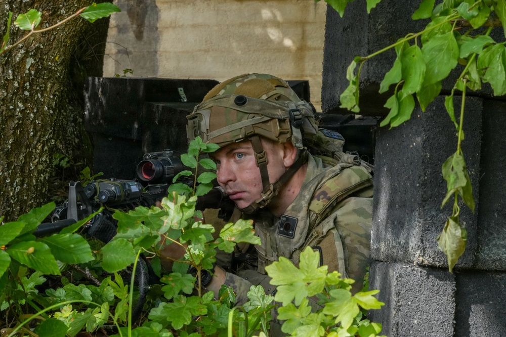 A soldier in full combat gear is positioned in concealment between a tree and a concrete block wall, aiming a rifle equipped with an optical scope and laser/light device. The soldier is wearing a multicam camouflage uniform and a tactical helmet with mounted accessories. The setting appears to be a military training exercise or operational environment with urban and natural elements present.