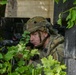 A soldier in full combat gear is positioned in concealment between a tree and a concrete block wall, aiming a rifle equipped with an optical scope and laser/light device. The soldier is wearing a multicam camouflage uniform and a tactical helmet with mounted accessories. The setting appears to be a military training exercise or operational environment with urban and natural elements present.