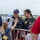 Two female skydivers in dark jumpsuits with American flag patches are greeting spectators at what appears to be an airshow. One skydiver wearing a blue Nike cap is receiving a high-five from a young blonde child over a metal barrier. The atmosphere is celebratory and joyful, with VIP badge holders visible among the crowd.