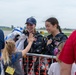 Two female skydivers in dark jumpsuits with American flag patches are greeting spectators at what appears to be an airshow. One skydiver wearing a blue Nike cap is receiving a high-five from a young blonde child over a metal barrier. The atmosphere is celebratory and joyful, with VIP badge holders visible among the crowd.