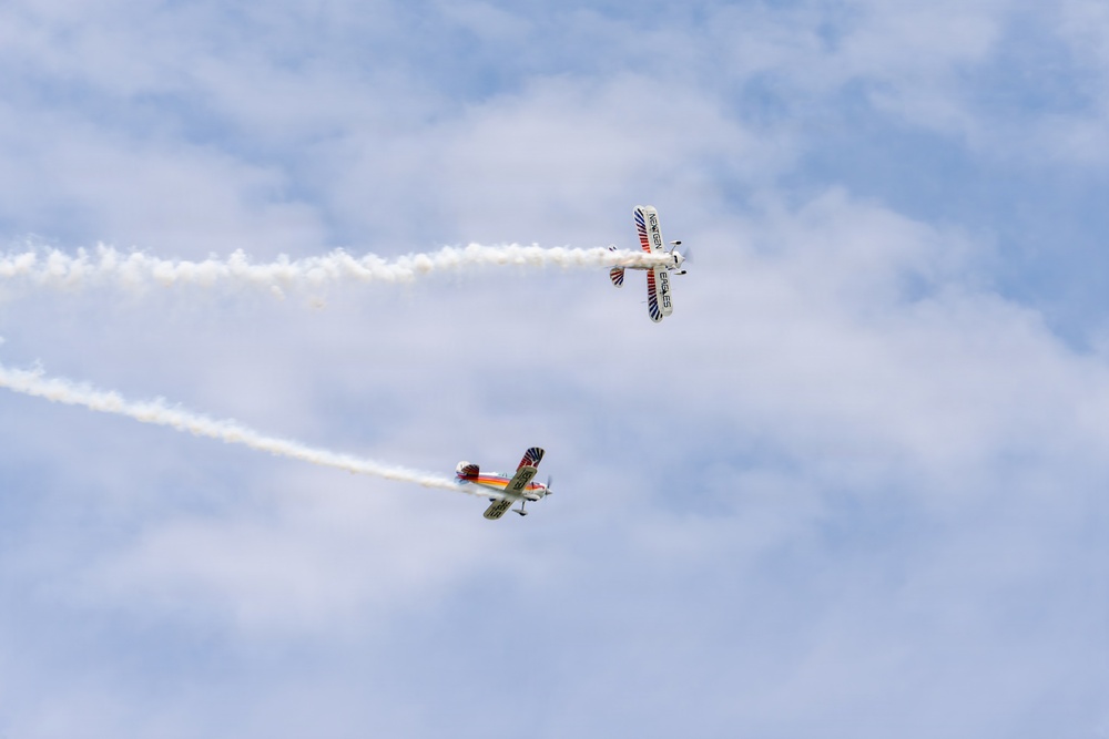 Two small aerobatic aircraft are performing a crossover maneuver at an airshow against a partly cloudy blue sky. Both aircraft are trailing white smoke streams that form an X-pattern as they pass near each other. The upper aircraft appears to be a biplane while the lower one is a monoplane, both displaying colorful liveries.