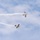 Two small aerobatic aircraft are performing a crossover maneuver at an airshow against a partly cloudy blue sky. Both aircraft are trailing white smoke streams that form an X-pattern as they pass near each other. The upper aircraft appears to be a biplane while the lower one is a monoplane, both displaying colorful liveries.