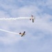 Two small aerobatic aircraft are performing a crossover maneuver at an airshow against a partly cloudy blue sky. Both aircraft are trailing white smoke streams that form an X-pattern as they pass near each other. The upper aircraft appears to be a biplane while the lower one is a monoplane, both displaying colorful liveries.